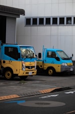 Two small trucks are parked side by side in front of a white building with several small windows. The trucks are painted in vibrant blue and yellow colors. The area in front of the trucks includes a concrete surface with road markings and a curb.