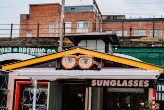 A vibrant storefront features a sign with large, stylized glasses and eyes above the entrance. The sign displays the word 'SUNGLASSES' in bold letters. The building's exterior includes a mix of brick and industrial-style elements, with an overpass or railway bridge in the background. A colorful collection of urban elements frame the shop, adding to the whimsical atmosphere.