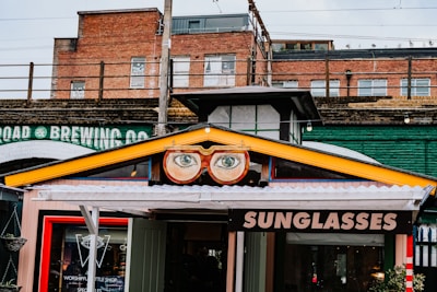 A vibrant storefront features a sign with large, stylized glasses and eyes above the entrance. The sign displays the word 'SUNGLASSES' in bold letters. The building's exterior includes a mix of brick and industrial-style elements, with an overpass or railway bridge in the background. A colorful collection of urban elements frame the shop, adding to the whimsical atmosphere.