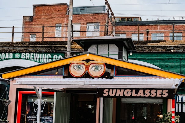 A vibrant storefront features a sign with large, stylized glasses and eyes above the entrance. The sign displays the word 'SUNGLASSES' in bold letters. The building's exterior includes a mix of brick and industrial-style elements, with an overpass or railway bridge in the background. A colorful collection of urban elements frame the shop, adding to the whimsical atmosphere.