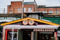 A vibrant storefront features a sign with large, stylized glasses and eyes above the entrance. The sign displays the word 'SUNGLASSES' in bold letters. The building's exterior includes a mix of brick and industrial-style elements, with an overpass or railway bridge in the background. A colorful collection of urban elements frame the shop, adding to the whimsical atmosphere.