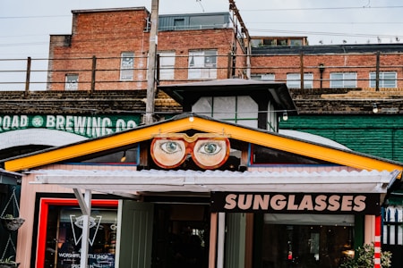 A vibrant storefront features a sign with large, stylized glasses and eyes above the entrance. The sign displays the word 'SUNGLASSES' in bold letters. The building's exterior includes a mix of brick and industrial-style elements, with an overpass or railway bridge in the background. A colorful collection of urban elements frame the shop, adding to the whimsical atmosphere.