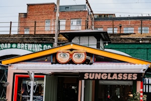A vibrant storefront features a sign with large, stylized glasses and eyes above the entrance. The sign displays the word 'SUNGLASSES' in bold letters. The building's exterior includes a mix of brick and industrial-style elements, with an overpass or railway bridge in the background. A colorful collection of urban elements frame the shop, adding to the whimsical atmosphere.