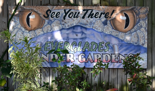 A colorful sign featuring a detailed illustration of a reptile's eyes, possibly a snake, with the words 'See You There!' in black at the top. Below, the text 'EVERGLADES WONDER GARDENS' is prominently displayed. The sign is partially obscured by green foliage, and is mounted on a wooden fence.
