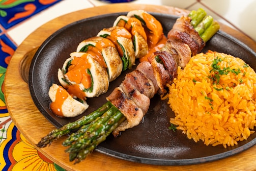 A plate containing rolled chicken breast slices drizzled with a reddish-orange sauce, accompanied by bacon-wrapped asparagus spears and a serving of yellow rice garnished with herbs. The plate is set on a table with colorful, patterned tiles.