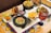 A colorful Mexican-themed table setting with a variety of dishes including guacamole in a stone bowl, a plate of tortilla chips, and a bowl of mixed vegetables such as cucumbers, carrots, and celery. There are two vibrant beverages, one in a large cup with a rim of spices and garnished with fruits. The table features decorative tiles with intricate patterns.