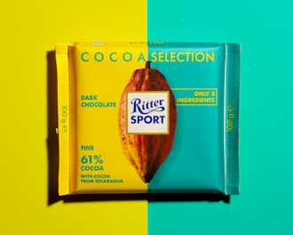 A chocolate bar wrapper divided into two contrasting backgrounds, yellow on the left and turquoise on the right. The wrapper features a cocoa pod illustration and text indicating it's a dark chocolate product with 61% cocoa from Nicaragua, highlighting its simplicity with only three ingredients.