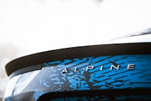 Close-up of a car's rear section displaying the word 'ALPINE' in metallic letters. The surface features a striking blue and black geometric pattern.