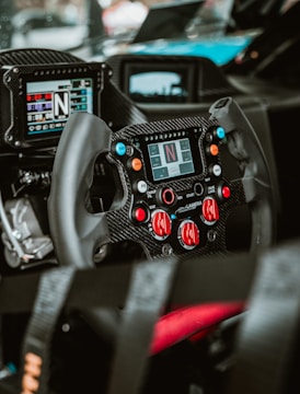 A close-up view of a high-tech racing car steering wheel with multiple buttons and controls. The dashboard features digital displays, and the steering wheel is made of carbon fiber, suggesting its use in a professional racing environment.