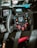 A close-up view of a high-tech racing car steering wheel with multiple buttons and controls. The dashboard features digital displays, and the steering wheel is made of carbon fiber, suggesting its use in a professional racing environment.