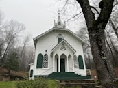 A white, classic-style church with green accents positioned in a serene forest environment. The church has a steeple, arched windows, and a central entrance with steps leading up. The surrounding trees are barren, indicating a winter or late fall setting.