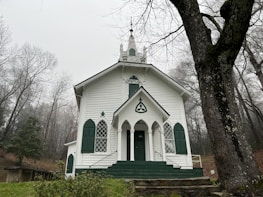 A white, classic-style church with green accents positioned in a serene forest environment. The church has a steeple, arched windows, and a central entrance with steps leading up. The surrounding trees are barren, indicating a winter or late fall setting.