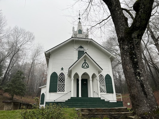 A white, classic-style church with green accents positioned in a serene forest environment. The church has a steeple, arched windows, and a central entrance with steps leading up. The surrounding trees are barren, indicating a winter or late fall setting.
