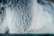 A close-up view of a textured and sculpted iceberg surface with intricate patterns and grooves, reflecting sunlight. The iceberg exhibits a mix of white and light blue hues, with darker blue water at its base.