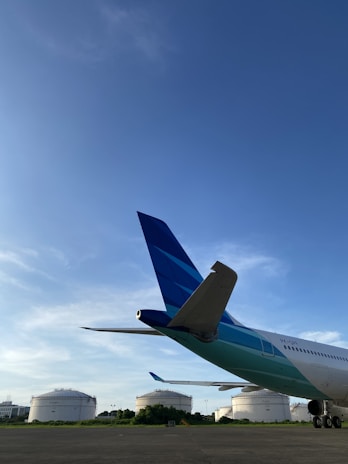 A commercial airplane is parked on a tarmac with its rear and tail prominently visible. In the background, there are large white fuel storage tanks surrounded by greenery. The sky is clear and blue, indicating good weather conditions.