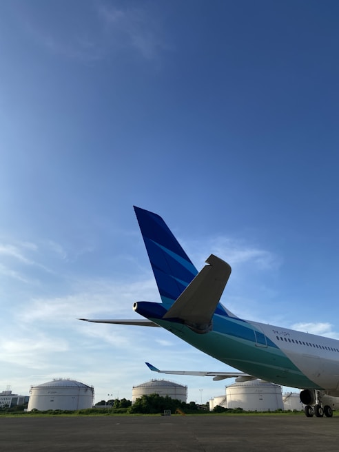 A commercial airplane is parked on a tarmac with its rear and tail prominently visible. In the background, there are large white fuel storage tanks surrounded by greenery. The sky is clear and blue, indicating good weather conditions.