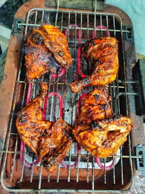 Four pieces of grilled chicken, perfectly cooked and charred, sit on a metal barbecue grill with visible smoke rising. The chicken appears to be seasoned with a rich, savory marinade, giving it a glistening and appetizing look.