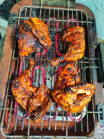 Four pieces of grilled chicken, perfectly cooked and charred, sit on a metal barbecue grill with visible smoke rising. The chicken appears to be seasoned with a rich, savory marinade, giving it a glistening and appetizing look.