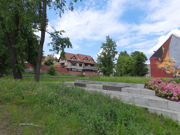 A semi-urban park area with seating and lush greenery, featuring vibrant flowers and large trees. In the background, traditional-style buildings with red roofs are visible, against a sky filled with scattered clouds. A mural decorates the side of one building.