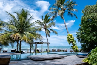 A serene beach scene with tall palm trees swaying gently in the breeze, overlooking an infinity pool that seamlessly merges with the ocean in the background. A wooden deck and lounge chairs suggest a relaxing atmosphere under the bright blue sky dotted with fluffy white clouds.