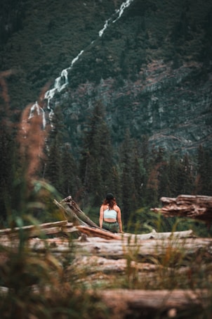 A person wearing a white top and green pants stands among fallen logs in a forested area. The background features dense evergreen trees on a hillside and a narrow, cascading waterfall. The image conveys a sense of tranquility and adventure.