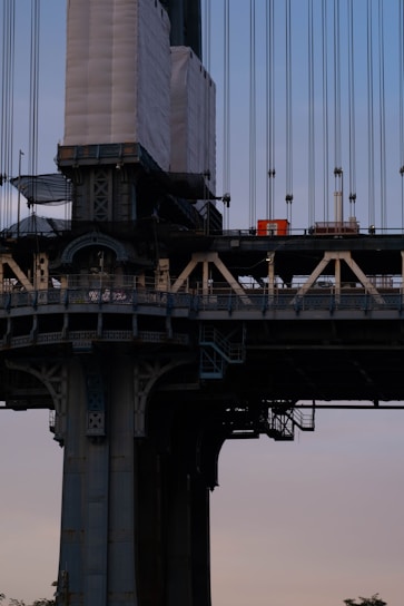 A close-up view of a bridge structure with various architectural details. The image captures the intricate lattice patterns and robust construction elements. Scaffolding and wrapped materials suggest ongoing maintenance or construction work.