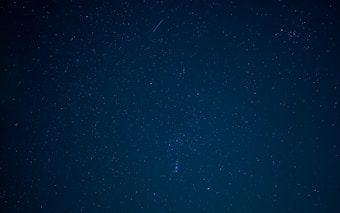 A night sky filled with numerous stars, creating a sparkling and serene celestial scene. A faint meteor streaks across the upper section of the image.