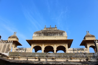 A traditional Indian architectural structure with ornate carvings and domed roofs set against a clear blue sky. The design features intricate latticework and decorative elements typical of historic buildings.