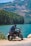 A person with a backpack is sitting on a motorcycle, overlooking a serene lake surrounded by mountains and dense forests. The scene is lit by bright daylight, creating a peaceful and adventurous ambiance.