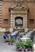 An outdoor café setting with multiple tables and chairs arranged on a cobblestone surface. The backdrop features an ornate stone façade with Latin inscriptions and a sculptural relief. Several tables have blue cloths draped over some chairs, and potted plants with green leaves and red flowers are placed in the foreground.