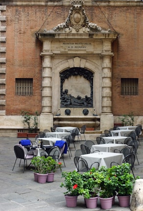 An outdoor café setting with multiple tables and chairs arranged on a cobblestone surface. The backdrop features an ornate stone façade with Latin inscriptions and a sculptural relief. Several tables have blue cloths draped over some chairs, and potted plants with green leaves and red flowers are placed in the foreground.