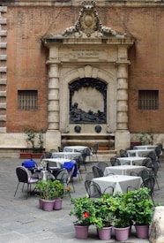 An outdoor caf&eacute; setting with multiple tables and chairs arranged on a cobblestone surface. The backdrop features an ornate stone fa&ccedil;ade with Latin inscriptions and a sculptural relief. Several tables have blue cloths draped over some chairs, and potted plants with green leaves and red flowers are placed in the foreground.