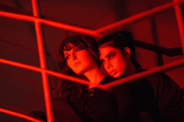 Two women are sitting closely together, illuminated by intense red lighting. They appear contemplative, surrounded by metal railings that intersect the composition, adding a geometric element to the scene.