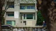 A modern building with a minimalist design featuring large windows and a sign that reads 'style and co'. The facade is predominantly gray, complemented by green-tinted windows. The setting is suburban, with trees and a hedge lining the front of the property. Cars are visible on the street in front.