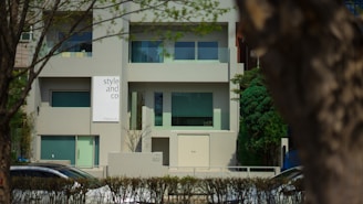 A modern building with a minimalist design featuring large windows and a sign that reads 'style and co'. The facade is predominantly gray, complemented by green-tinted windows. The setting is suburban, with trees and a hedge lining the front of the property. Cars are visible on the street in front.