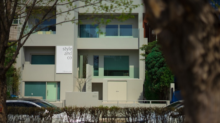 A modern building with a minimalist design featuring large windows and a sign that reads 'style and co'. The facade is predominantly gray, complemented by green-tinted windows. The setting is suburban, with trees and a hedge lining the front of the property. Cars are visible on the street in front.