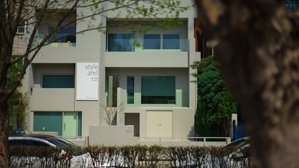 A modern building with a minimalist design featuring large windows and a sign that reads 'style and co'. The facade is predominantly gray, complemented by green-tinted windows. The setting is suburban, with trees and a hedge lining the front of the property. Cars are visible on the street in front.