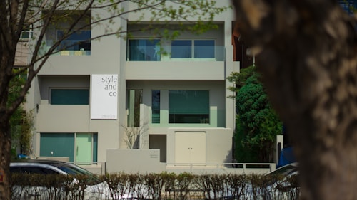 A modern building with a minimalist design featuring large windows and a sign that reads 'style and co'. The facade is predominantly gray, complemented by green-tinted windows. The setting is suburban, with trees and a hedge lining the front of the property. Cars are visible on the street in front.