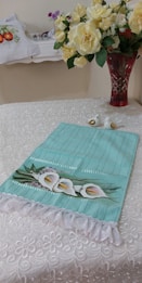 A decorative table setting featuring a lace-patterned tablecloth with a teal towel adorned with white calla lilies embroidery. A red glass vase with large yellow and white flowers is placed on the table, creating a centerpiece. A small ceramic bird ornament sits nearby, adding a delicate touch.