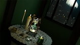 A dimly lit room with a small round table draped with a lace tablecloth. On the table, there is an antique hourglass, tarot cards, a burning candle, and a stack of cards. A window behind reveals a night sky with a full moon and tree silhouettes.
