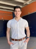 A man is standing in a brightly lit area, posing with hands in his pockets. He is wearing a white short-sleeve collared shirt and light beige pants with a brown belt. The background consists of an industrial setting, with a combination of orange and dark blue walls, and a white ceiling structure with exposed beams. There are numbered signs on the walls and a 'FUMAR' sign indicating a smoking area.
