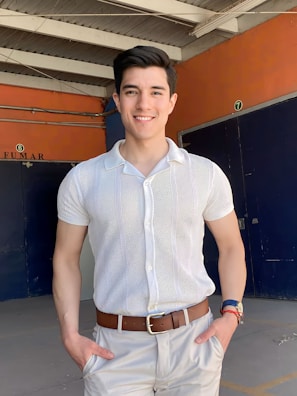 A man is standing in a brightly lit area, posing with hands in his pockets. He is wearing a white short-sleeve collared shirt and light beige pants with a brown belt. The background consists of an industrial setting, with a combination of orange and dark blue walls, and a white ceiling structure with exposed beams. There are numbered signs on the walls and a 'FUMAR' sign indicating a smoking area.