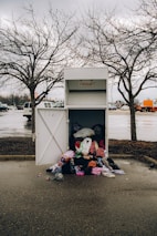 An overflowing donation bin with clothes spilling out onto the wet pavement. It is placed in a parking lot under overcast skies, surrounded by barren trees and parked vehicles.