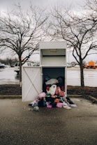 An overflowing donation bin with clothes spilling out onto the wet pavement. It is placed in a parking lot under overcast skies, surrounded by barren trees and parked vehicles.