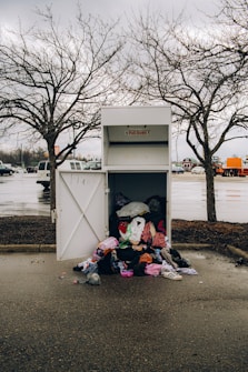 An overflowing donation bin with clothes spilling out onto the wet pavement. It is placed in a parking lot under overcast skies, surrounded by barren trees and parked vehicles.