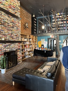 A cozy bookstore interior with tall shelves filled with books, a brick wall, and wooden floors. The space has a black leather sofa and a fireplace. There are hanging lights and a plant placed near the fireplace. A table displays various items and a wooden ladder leans against the shelves. An artistic tree structure is part of the decor.