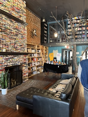 A cozy bookstore interior with tall shelves filled with books, a brick wall, and wooden floors. The space has a black leather sofa and a fireplace. There are hanging lights and a plant placed near the fireplace. A table displays various items and a wooden ladder leans against the shelves. An artistic tree structure is part of the decor.