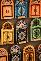 Colorful decorative items resembling miniature window frames are displayed. Each piece is intricately designed with various patterns, featuring bold colors like orange, blue, yellow, and green. Some have small wooden doors with detailed carvings and floral motifs.