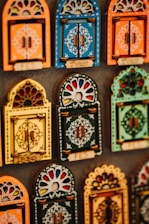 Colorful decorative items resembling miniature window frames are displayed. Each piece is intricately designed with various patterns, featuring bold colors like orange, blue, yellow, and green. Some have small wooden doors with detailed carvings and floral motifs.