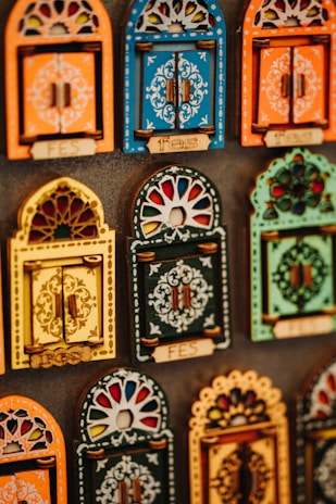 Colorful decorative items resembling miniature window frames are displayed. Each piece is intricately designed with various patterns, featuring bold colors like orange, blue, yellow, and green. Some have small wooden doors with detailed carvings and floral motifs.